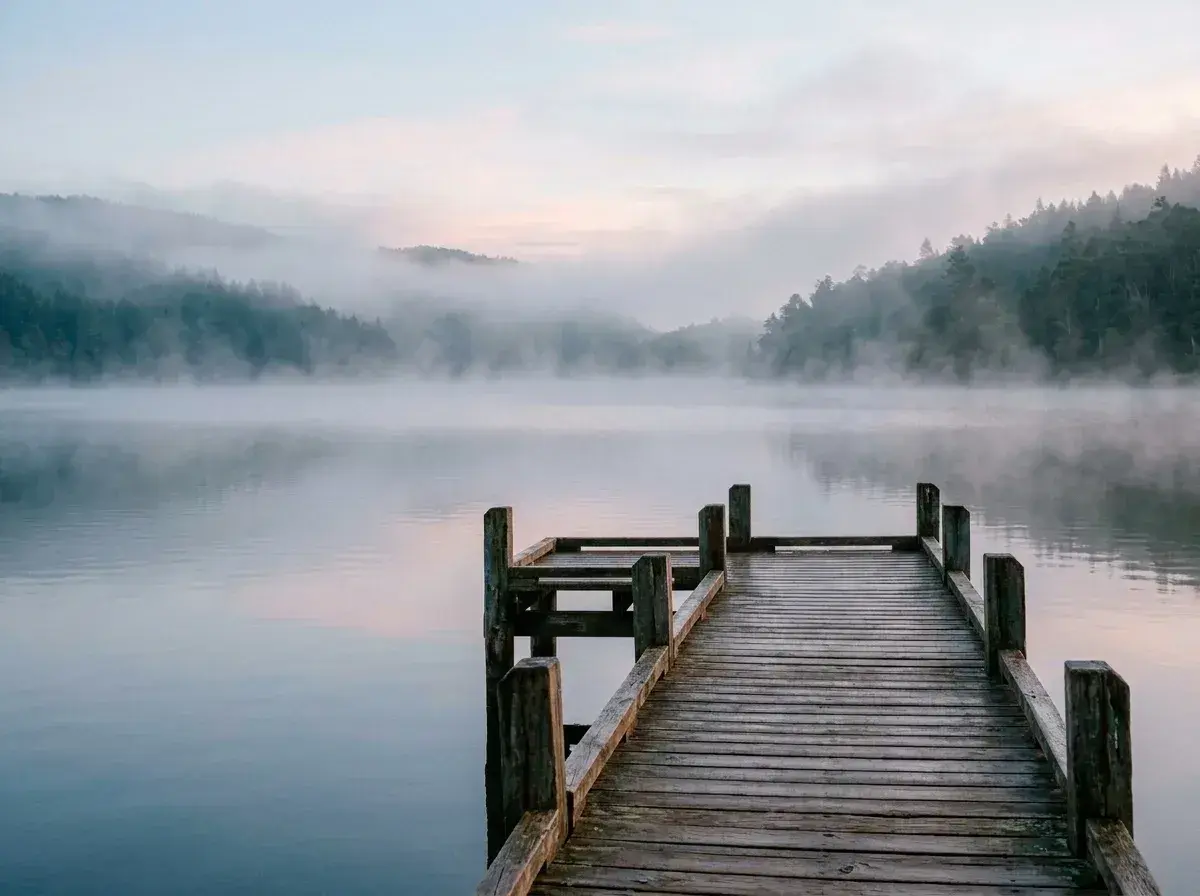 An empty wooden pier extends into a calm, misty lake surrounded by hills covered in trees.