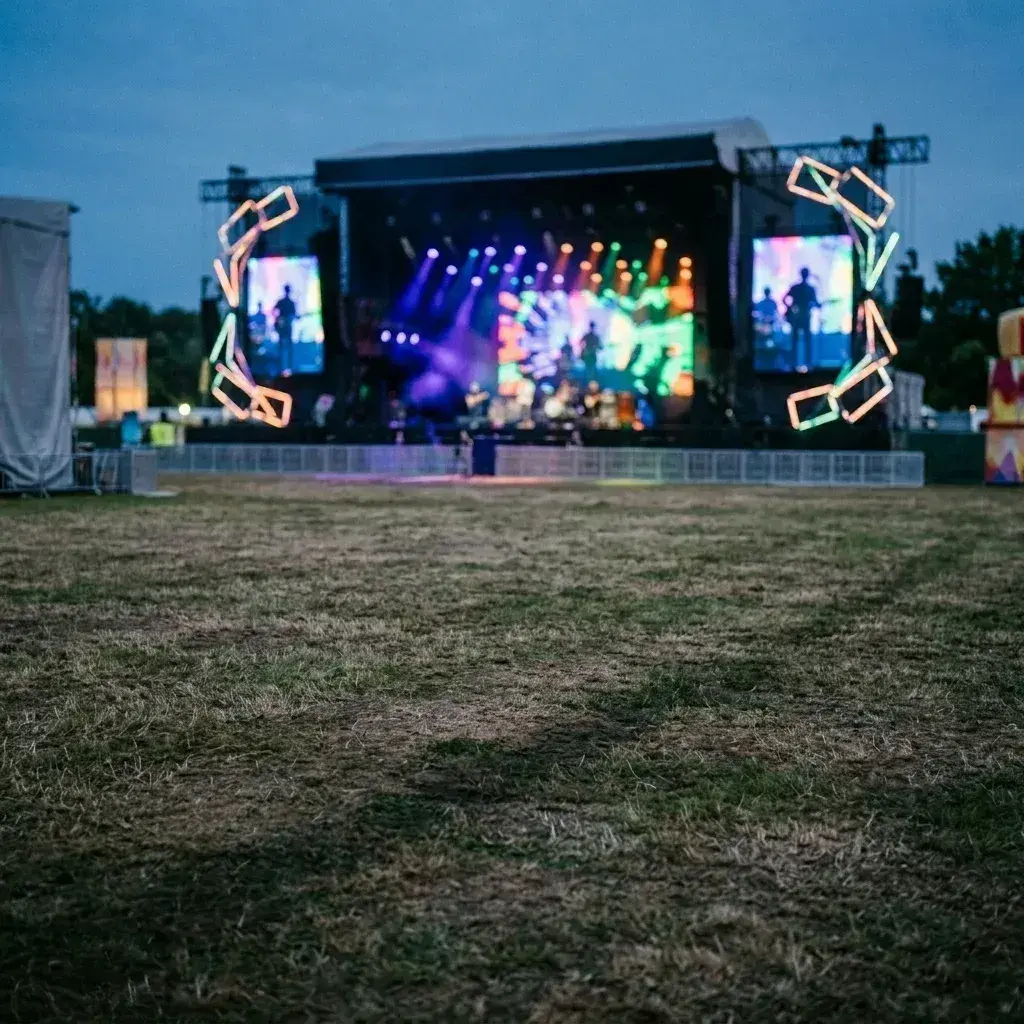 An empty grassy field at dusk facing a brightly lit concert stage with no people visible.