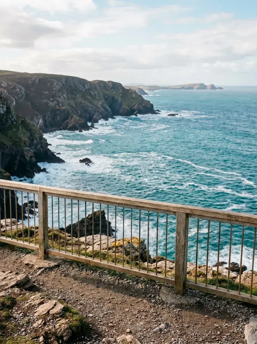 A wooden railing stands on a rocky path overlooking a blue ocean with waves crashing against steep cliffs.