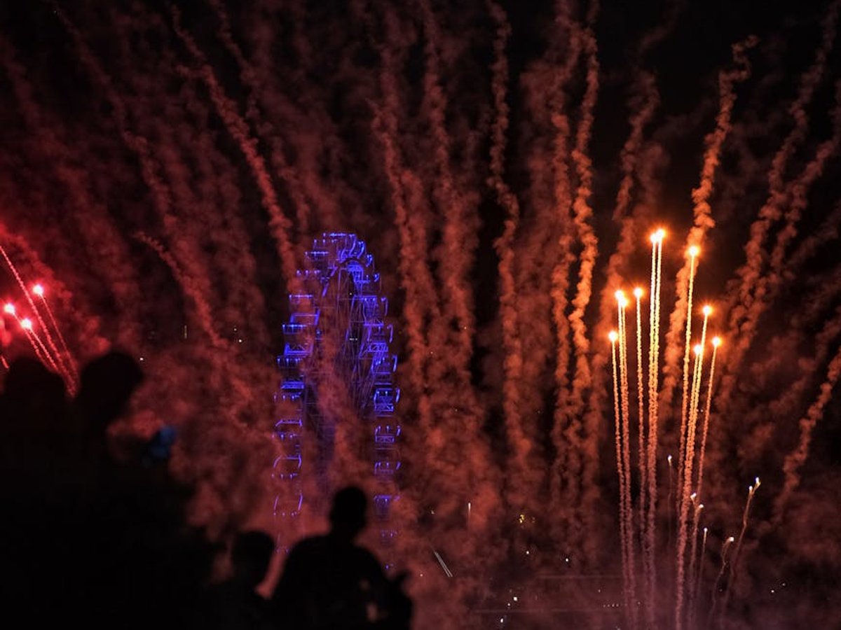 Fireworks illuminate a Ferris wheel at night, people silhouettes watching the celebration. - before AI removal